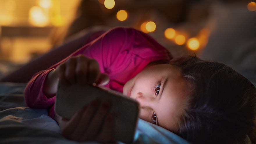 Small girl indoors on bed at night, using cell phone.