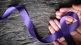 Top view of male hand holding purple cancer ribbon on wood background